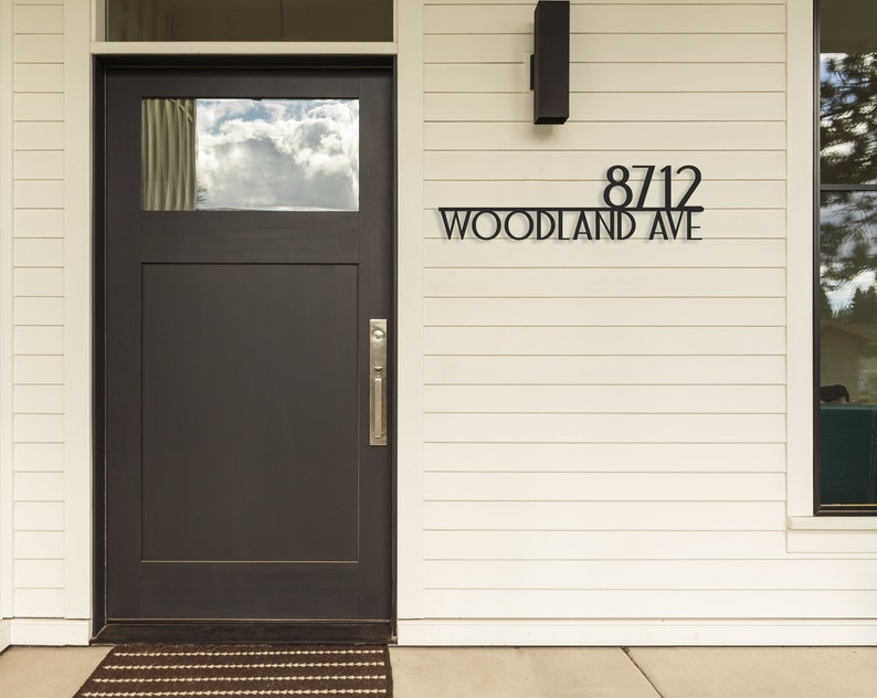 May include: A black metal house number sign with the number 8712 and the street name "WOODLAND AVE" on a white house exterior.