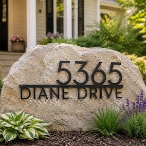 May include: A large, light-colored natural boulder featuring a black metal address sign mounted on its front. The sign displays the number 5365 in large digits above a horizontal bar, with the text DIANE DRIVE in capital letters below the bar.