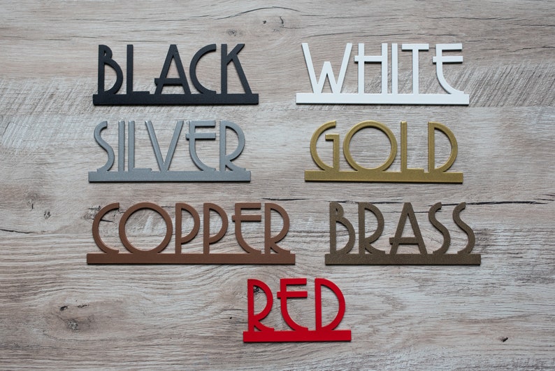 May include: A collection of metal word cutouts in various colors, including black, white, silver, gold, copper, brass, and red. The words are arranged in a row on a wooden surface.