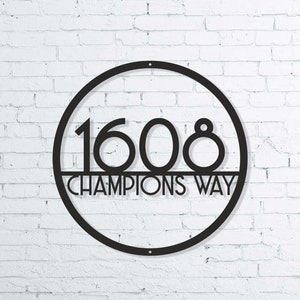 Puede incluir: Letrero de n&uacute;mero de casa de metal negro con un marco circular y los n&uacute;meros 1608 y el texto "CHAMPIONS WAY" en un fondo de pared de ladrillo blanco.