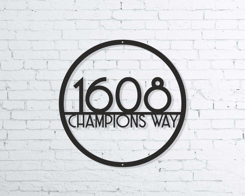 Puede incluir: Letrero de n&uacute;mero de casa de metal negro con un marco circular y los n&uacute;meros 1608 y el texto "CHAMPIONS WAY" en un fondo de pared de ladrillo blanco.