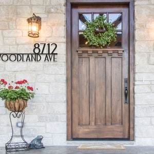 May include: Black metal house number sign with the number 8712 and the text "Woodland Ave" on a stone wall with a wooden door and a wreath.