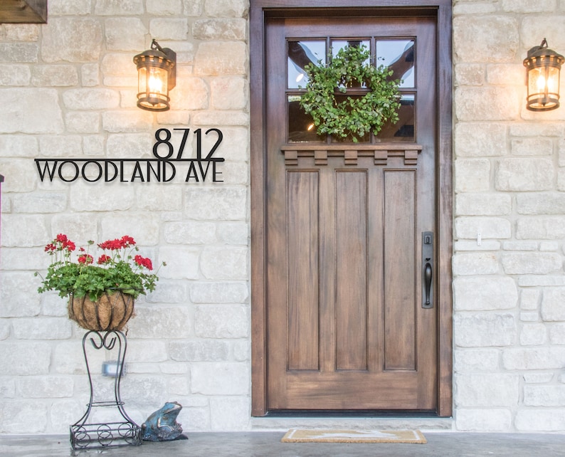 May include: Black metal house number sign with the number 8712 and the text "Woodland Ave" on a stone wall with a wooden door and a wreath.