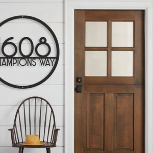 Puede incluir: Placa de n&uacute;mero de casa de metal negro con un marco circular y el n&uacute;mero 1608 y el texto "Champions Way" en una pared de madera blanca.