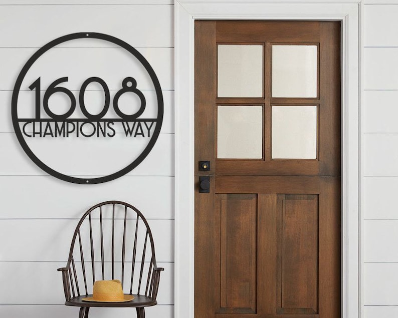 Puede incluir: Placa de n&uacute;mero de casa de metal negro con un marco circular y el n&uacute;mero 1608 y el texto "Champions Way" en una pared de madera blanca.