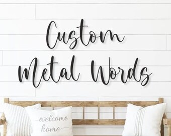 以下が含まれることがあります： 「Custom Metal Words」というテキストが筆記体で書かれたカスタムメタルワードサイン。サインは白い木製の壁に、木製のベンチと2つのクッションの上に吊り下げられています。