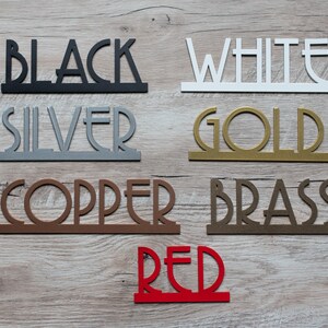 Puede incluir: Una colecci&oacute;n de recortes de palabras de metal en varios colores, incluyendo negro, blanco, plata, oro, cobre, lat&oacute;n y rojo. Las palabras est&aacute;n dispuestas en una fila sobre una superficie de madera.