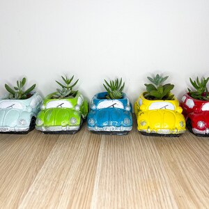 May include: Five miniature Volkswagen car planters in different colours, each with a small succulent plant growing inside. The planters are arranged in a row on a wooden surface.