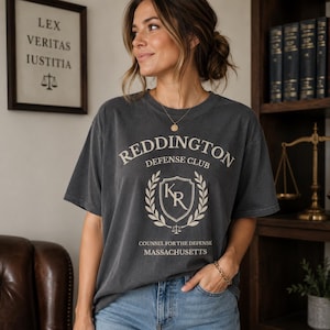 May include: A charcoal gray short-sleeve t-shirt featuring the text REDDINGTON DEFENSE CLUB, a shield emblem with the letters KR, and the words COUNSEL FOR THE DEFENSE MASSACHUSETTS printed in a light cream color on the front of the garment.