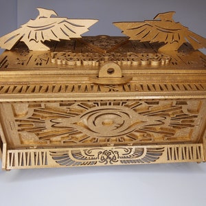 May include: A gold-colored, intricately carved box with an Egyptian theme. The box features detailed geometric patterns, hieroglyphs, and stylized bird figures. The lid has a latch and is topped with two bird-shaped ornaments.