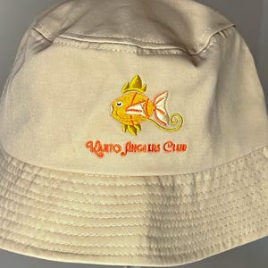 May include: Beige bucket hat with a stitched brim. The hat features an embroidered design of a yellow and orange fish with the text "Kanto Anglers Club" in orange thread. The hat is displayed on a stand.