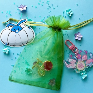 May include: A green tulle bag with a white and blue cartoon bunny sticker, a gold charm, and a clear plastic charm with a pink and blue cartoon character. The bag is on a light blue background with scattered glitter.