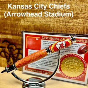 May include: A red, white, and gold pen with a Kansas City Chiefs logo and the words "Arrowhead Stadium" on the barrel. The pen is on a silver stand and has a certificate of authenticity.