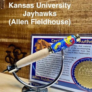 May include: A wooden pen with a blue and yellow Kansas Jayhawks themed pen top. The pen is on a silver stand and a certificate of authenticity is visible in the background. The certificate states that the material is sourced from the Allen Fieldhouse court, home of the University of Kansas Jayhawks basketball team.