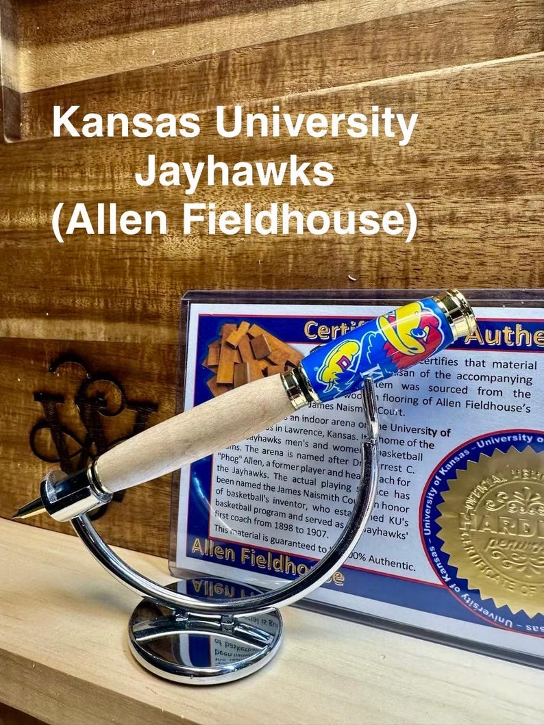 May include: A wooden pen with a blue and yellow Kansas Jayhawks themed pen top. The pen is on a silver stand and a certificate of authenticity is visible in the background. The certificate states that the material is sourced from the Allen Fieldhouse court, home of the University of Kansas Jayhawks basketball team.