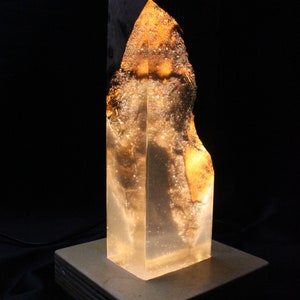 May include: A clear resin lamp with a wooden base. The resin has a golden hue and contains air bubbles. The lamp is lit and casting a warm glow.