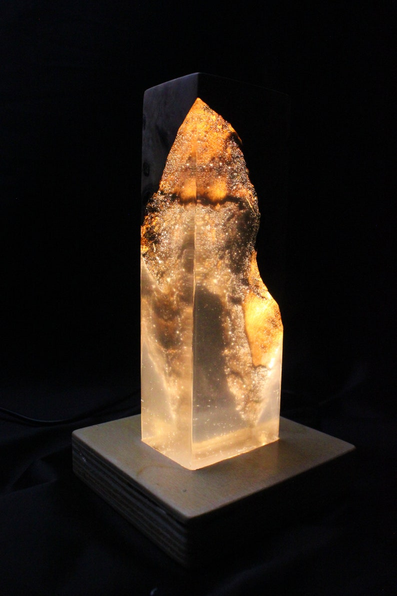 May include: A clear resin lamp with a wooden base. The resin has a golden hue and contains air bubbles. The lamp is lit and casting a warm glow.