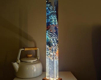 May include: A tall, clear resin lamp with a wooden base. The resin contains blue, green, and gold swirls. The lamp is lit and casting a warm glow.