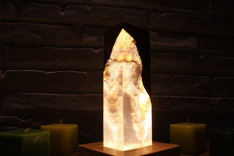 May include: A clear resin lamp with a wood base. The resin is shaped like a tall, thin rock with a light inside. The lamp is lit and casting a warm glow. There are three candles in the background.