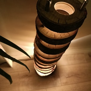 May include: A tall, cylindrical floor lamp made from stacked wooden rounds with a warm, glowing light emanating from the center. The lamp has a rustic, natural look and is perfect for adding a touch of warmth and ambiance to any room.