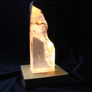 May include: A clear glass lamp with a golden resin base. The lamp has a unique, organic shape and is lit from within, creating a warm glow.