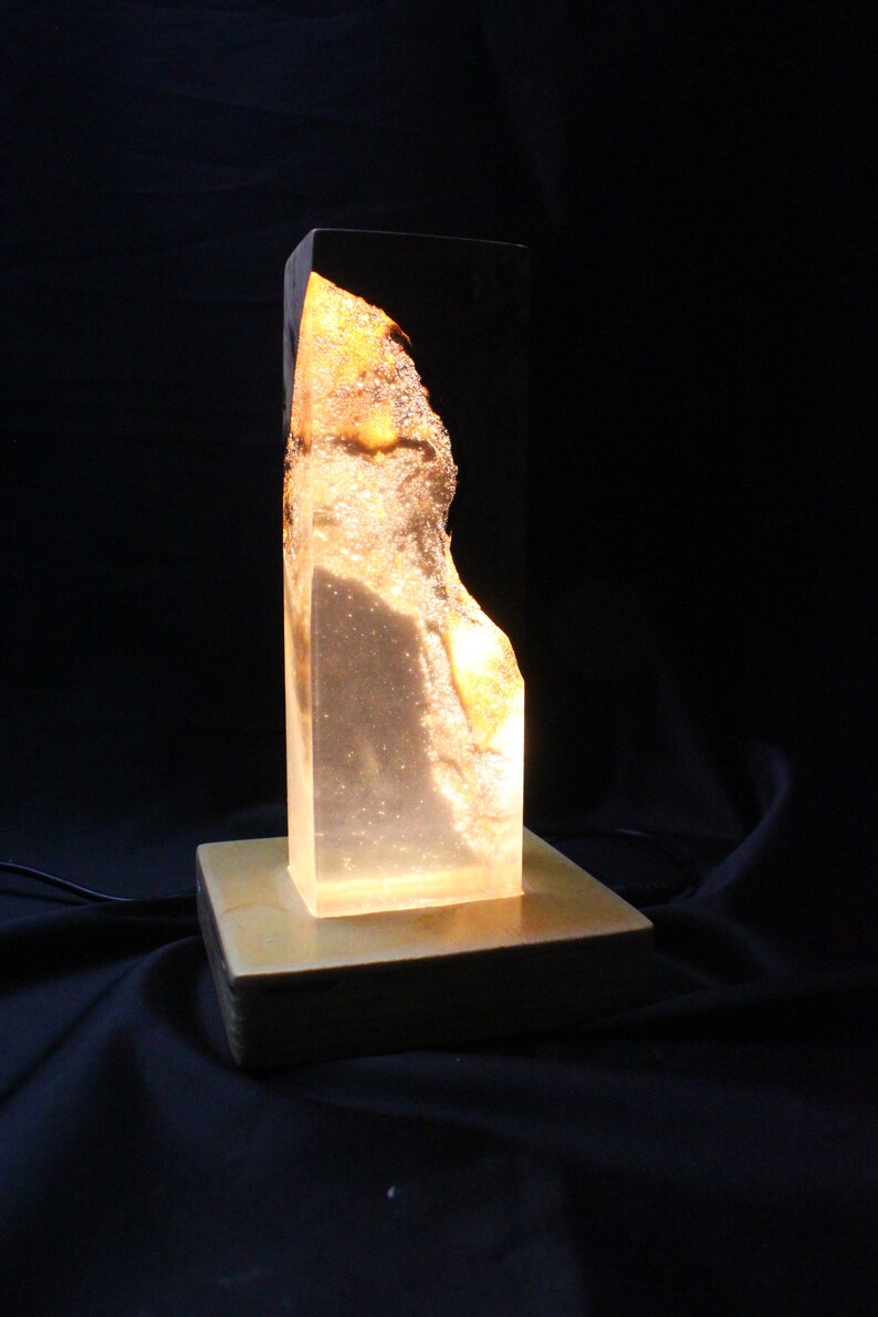 May include: A clear glass lamp with a golden resin base. The lamp has a unique, organic shape and is lit from within, creating a warm glow.