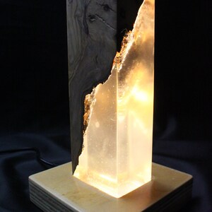 May include: A wooden and clear resin lamp with a warm, glowing light. The lamp has a natural wood base and a clear resin body with a wood grain pattern.