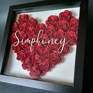 May include: A black framed shadow box with a white background. The box contains a heart shape made of red paper roses. The name "Simphoney" is written in white cursive lettering on the white background.