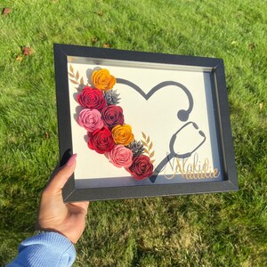 May include: A framed paper craft with a stethoscope and a heart shape surrounded by red, pink, and yellow paper flowers. The name "Natalie" is written in gold lettering at the bottom of the frame.