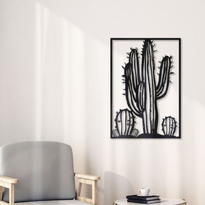 Puede incluir: Arte de pared de metal negro con un cactus estilizado de m&uacute;ltiples brazos. El cactus est&aacute; enmarcado en un marco cuadrado negro y se muestra en una pared blanca en un ambiente de sala de estar con un sill&oacute;n gris y una mesa auxiliar dorada.