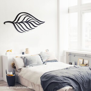 May include: Black metal leaf wall art hanging above a bed with white bedding and a gray throw blanket. The bedroom has a white wall and large windows.