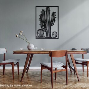 Puede incluir: Una escultura de pared de metal negro de una planta de cactus con tres cactus en total. La escultura est&aacute; colgada en una pared gris claro sobre una mesa de comedor de madera con cuatro sillas. La mesa est&aacute; puesta con un jarr&oacute;n de flores secas, vasos y un plato de fruta.