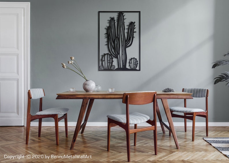 Puede incluir: Una escultura de pared de metal negro de una planta de cactus con tres cactus en total. La escultura est&aacute; colgada en una pared gris claro sobre una mesa de comedor de madera con cuatro sillas. La mesa est&aacute; puesta con un jarr&oacute;n de flores secas, vasos y un plato de fruta.