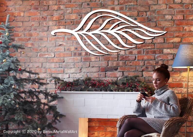 May include: White metal leaf wall art with a delicate design, hanging on a brick wall in a living room setting. A person is sitting in a wicker chair in front of a fireplace.