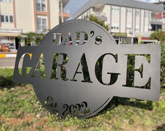 May include: A matte black metal sign featuring a circular and rectangular design with the cut-out text DAD'S GARAGE and Est. 2022. The sign has a smooth finish and bold, capitalized lettering that is clearly visible against the outdoor background.