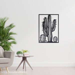 Puede incluir: Arte de pared de metal negro con una silueta de un cactus con un total de tres cactus. El arte est&aacute; enmarcado en un marco cuadrado negro y est&aacute; colgado en una pared blanca.