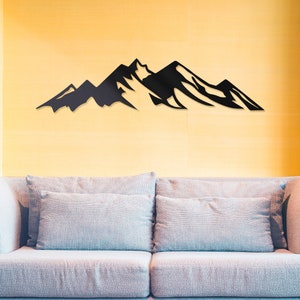 May include: A black metal mountain range wall decor silhouette mounted on a yellow wall above a light gray couch with two pillows.