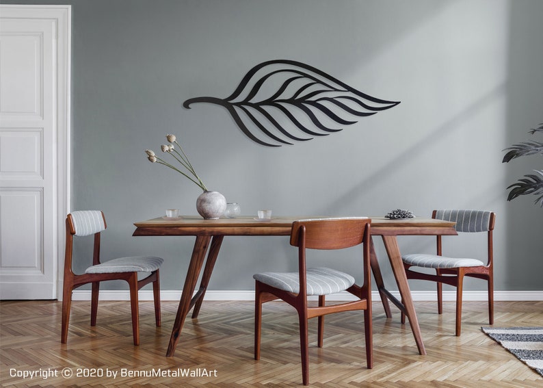 May include: A black metal leaf wall art sculpture hangs on a light gray wall above a wooden dining table with four chairs. The table has a light wood finish and the chairs have gray and white striped fabric seats.