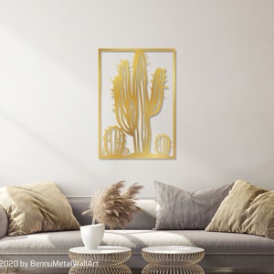 Puede incluir: Arte de pared de metal dorado con un dise&ntilde;o de cactus estilizado. El cactus est&aacute; enmarcado en un marco rectangular. La obra de arte se exhibe en una pared blanca sobre un sof&aacute; gris.