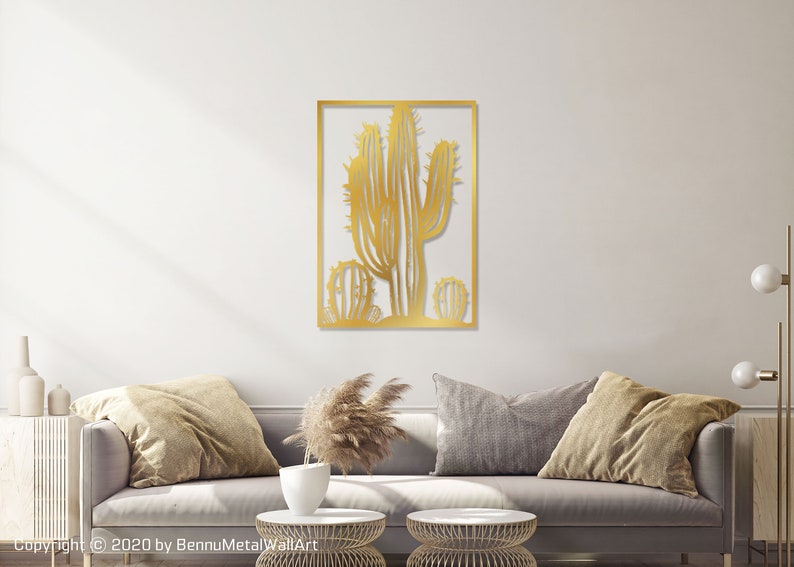 Puede incluir: Arte de pared de metal dorado con un dise&ntilde;o de cactus estilizado. El cactus est&aacute; enmarcado en un marco rectangular. La obra de arte se exhibe en una pared blanca sobre un sof&aacute; gris.