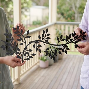May include: Black metal wall art depicting a stylized vine with leaves. The decorative piece is held by two people, showcasing its length and design. The art piece is suitable for indoor or outdoor decor.