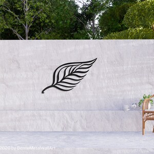 May include: A black metal leaf wall art sculpture mounted on a gray concrete wall. The sculpture is a stylized representation of a leaf with a central vein and multiple parallel lines extending from it. The wall art is displayed in a modern home setting with a wicker chair and a potted plant.
