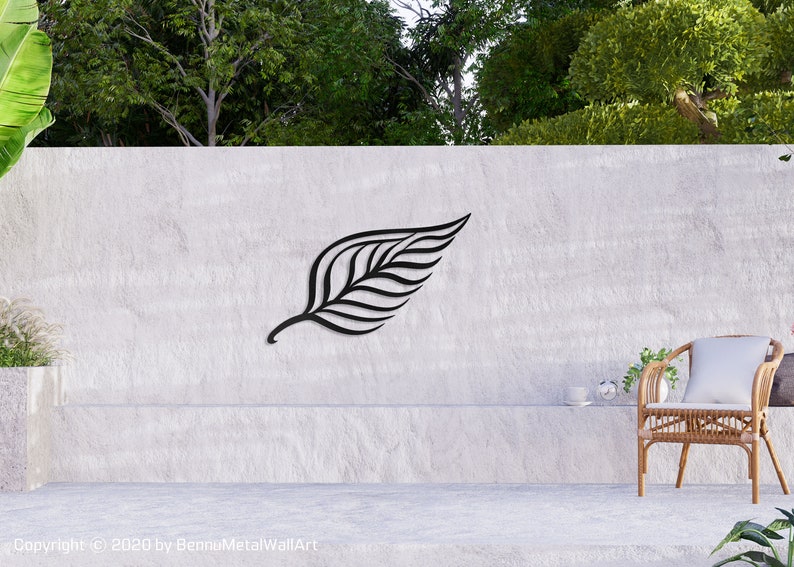 May include: A black metal leaf wall art sculpture mounted on a gray concrete wall. The sculpture is a stylized representation of a leaf with a central vein and multiple parallel lines extending from it. The wall art is displayed in a modern home setting with a wicker chair and a potted plant.