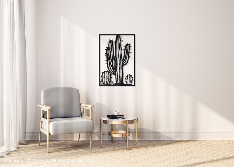Puede incluir: Arte de pared de metal negro con un cactus estilizado de m&uacute;ltiples brazos. El cactus est&aacute; enmarcado en un marco cuadrado negro y se muestra en una pared blanca en un ambiente de sala de estar con un sill&oacute;n gris y una mesa auxiliar dorada.