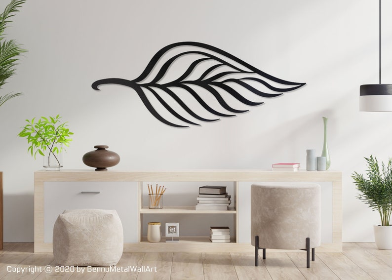 May include: Black metal wall art in the shape of a large leaf with a detailed design. The art is mounted on a white wall above a wooden shelf with books and decorative items.