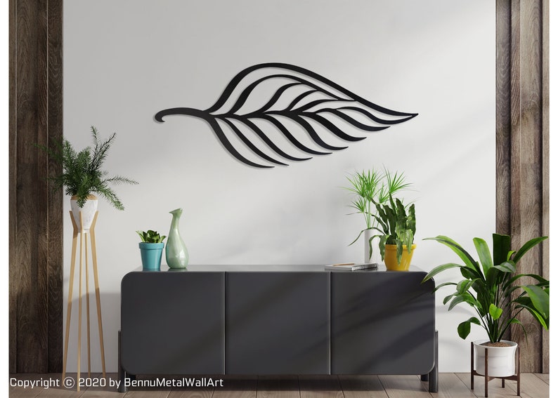 May include: Black metal leaf wall art with a modern design. The leaf is hanging on a white wall above a black dresser with three drawers. There are three potted plants on the dresser and a potted plant on a stand to the left of the dresser.