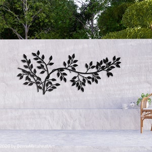 May include: Black metal wall art depicting a stylized branch with leaves. The artwork is mounted on a textured white wall, with a natural outdoor setting in the background. A wicker chair with a white cushion is on the right.