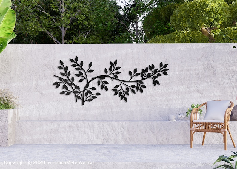 May include: Black metal wall art depicting a stylized branch with leaves. The artwork is mounted on a textured white wall, with a natural outdoor setting in the background. A wicker chair with a white cushion is on the right.