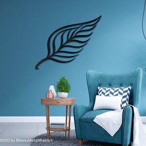 May include: A black metal leaf wall art decor hanging on a teal blue wall. A teal blue armchair with a white throw blanket and a patterned pillow sits in front of the wall art. A small wooden side table with a potted plant and decorative items sits next to the armchair.