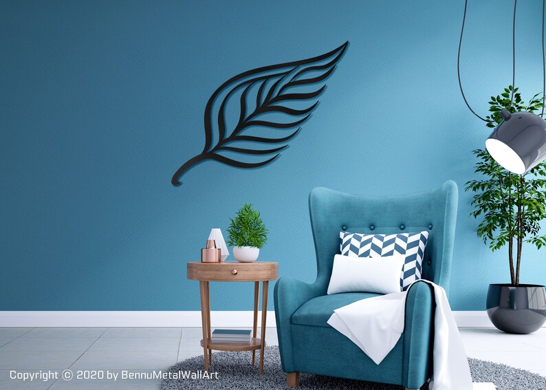 May include: A black metal leaf wall art decor hanging on a teal blue wall. A teal blue armchair with a white throw blanket and a patterned pillow sits in front of the wall art. A small wooden side table with a potted plant and decorative items sits next to the armchair.